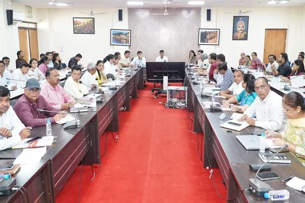 सभाकक्ष में यू आकार की मेजों पर बैठे अधिकारी दस्तावेजों और लैपटॉप के साथ बैठक करते हुए